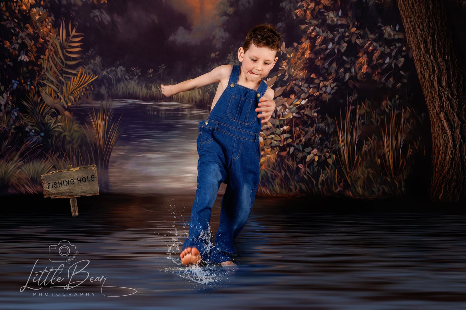 Young boy in blue denim overalls jumps and splashes in water against a dark forest backdrop.