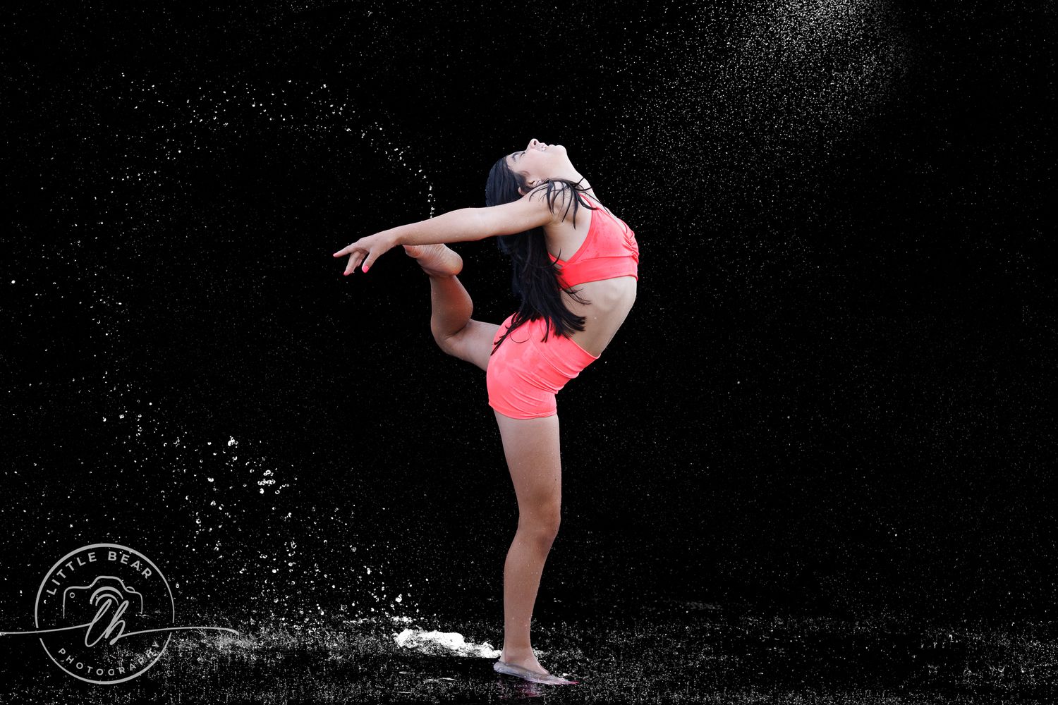 A dancer in pink athletic wear performs a graceful arabesque pose with water droplets splashing around her on black background.