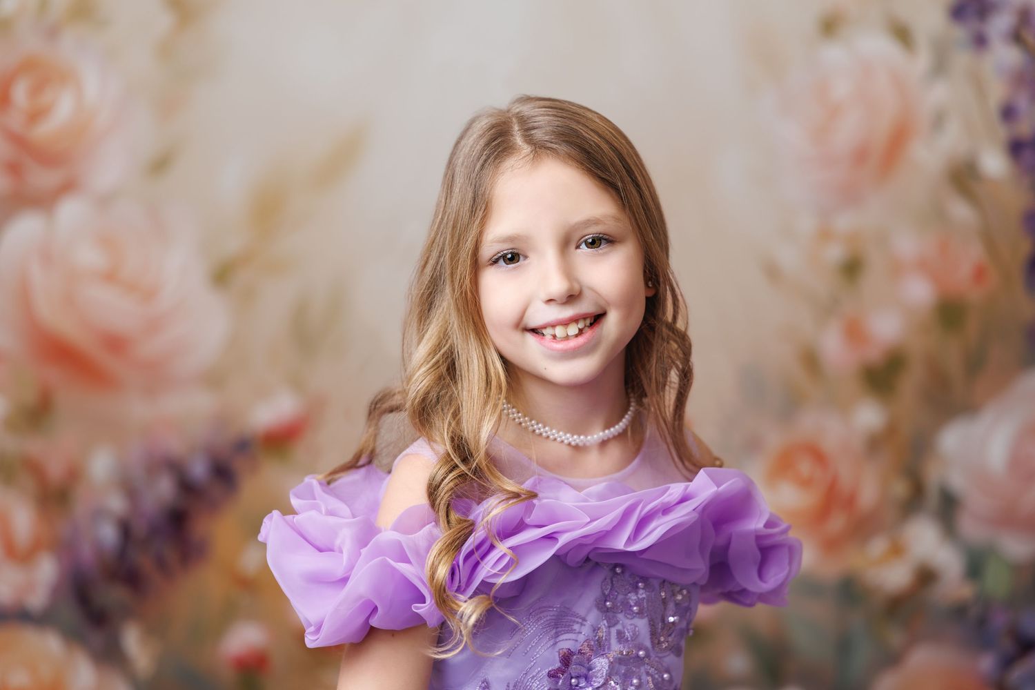 Smiling young girl in a purple ruffled dress posed in front of a soft floral painted photography backdrop.