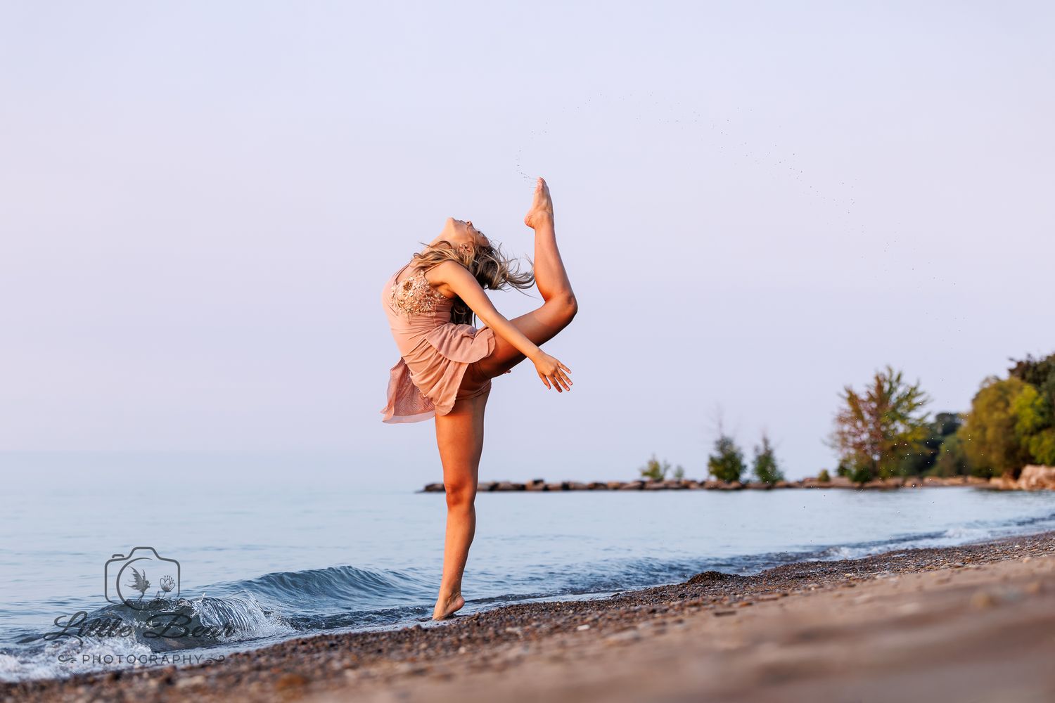 A graceful dancer performs a high kick on a sandy beach at sunset wearing a flowing pink dress.