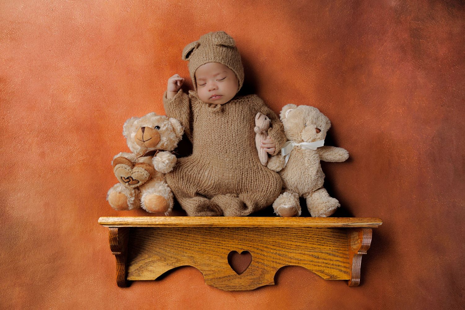Sleeping newborn in brown bear outfit rests on wooden shelf flanked by two teddy bears.