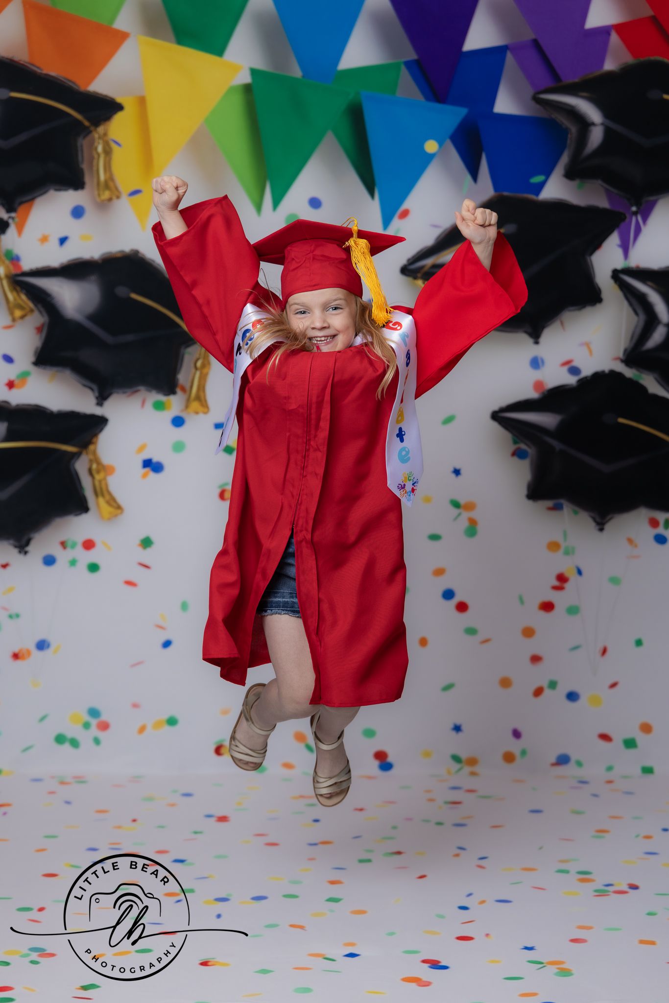 Young girl in red graduation cap and gown jumping joyfully with confetti and balloon decorations.