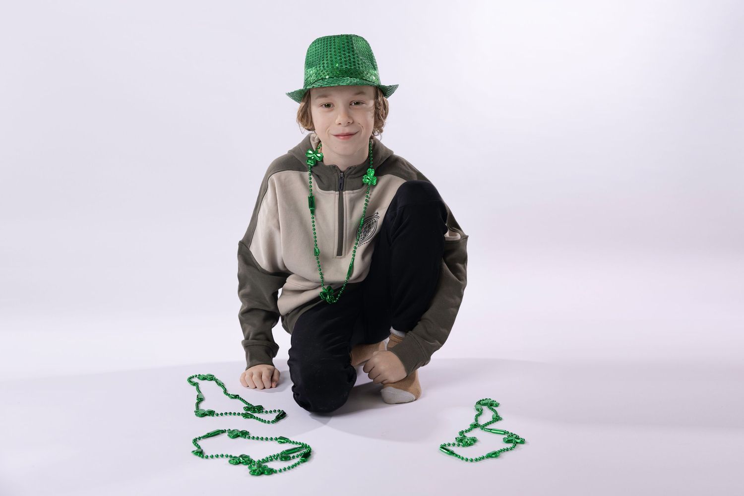 A young boy in a green hat crouches beside green St. Patrick's Day bead necklaces on a white background.