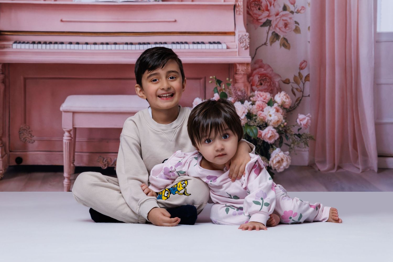 Young boy and toddler girl sit together smiling in front of pink piano in floral decorated studio.