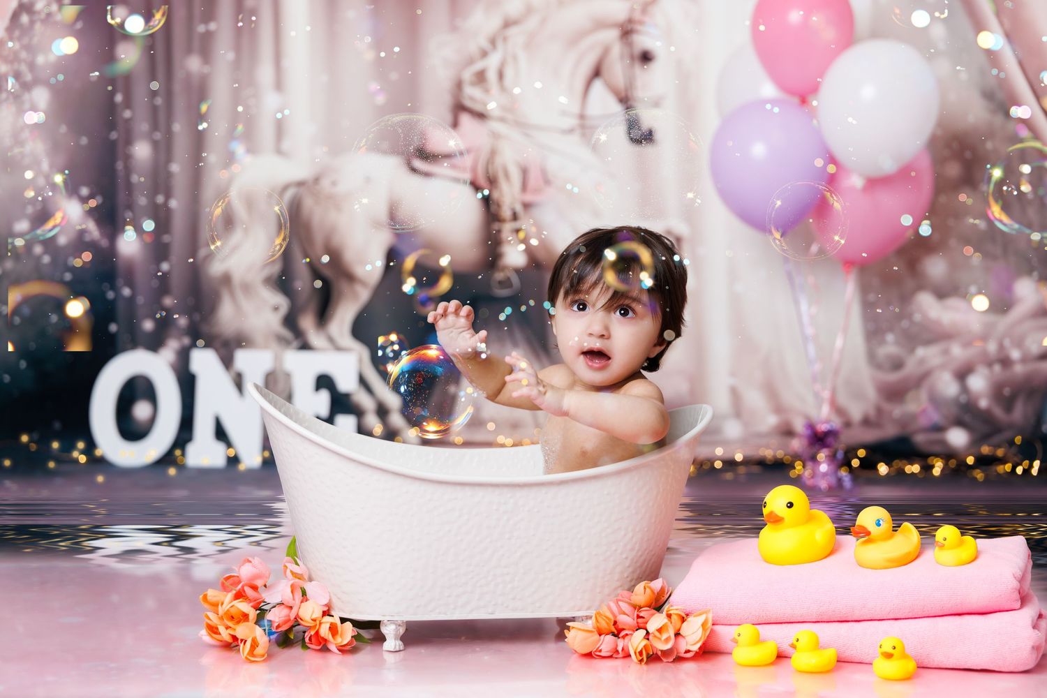 Baby sitting in a white clawfoot bathtub with rubber ducks and balloons during a first birthday cake smash photo session.