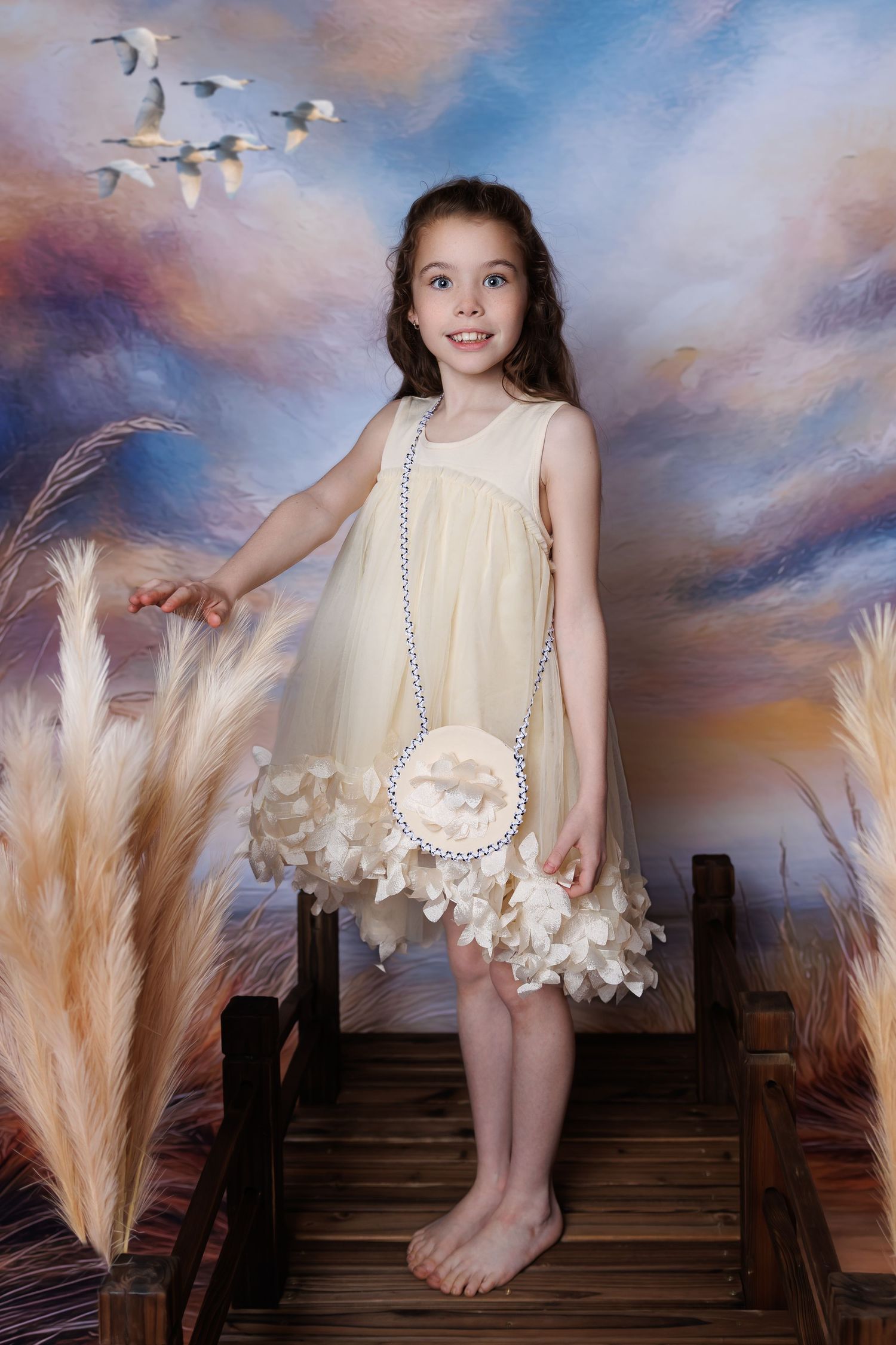 A smiling young girl in a cream floral dress stands barefoot on a wooden bridge with a painted sky backdrop.