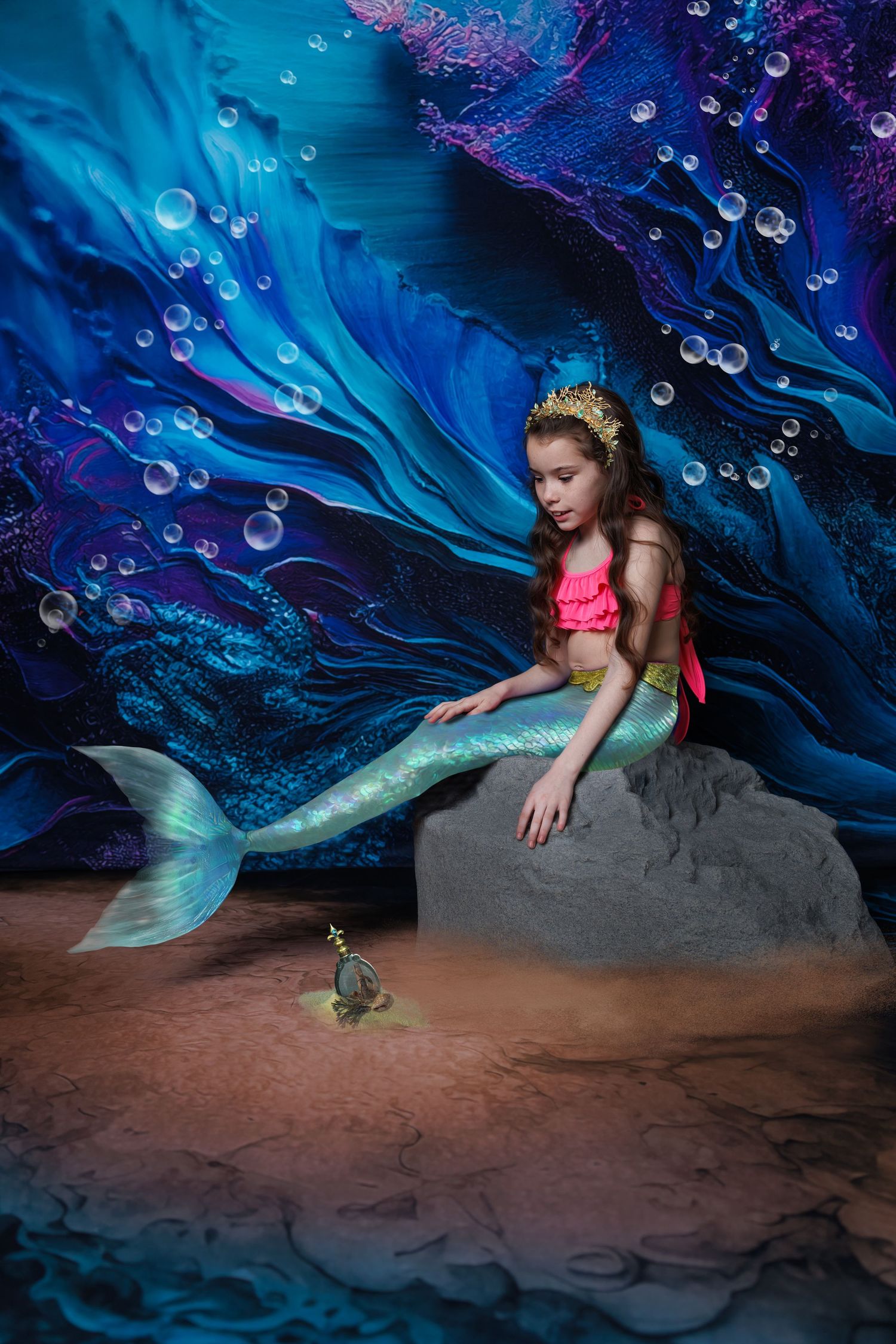 Girl in mermaid costume with gold crown sits on rock with bubbles and underwater ocean backdrop.