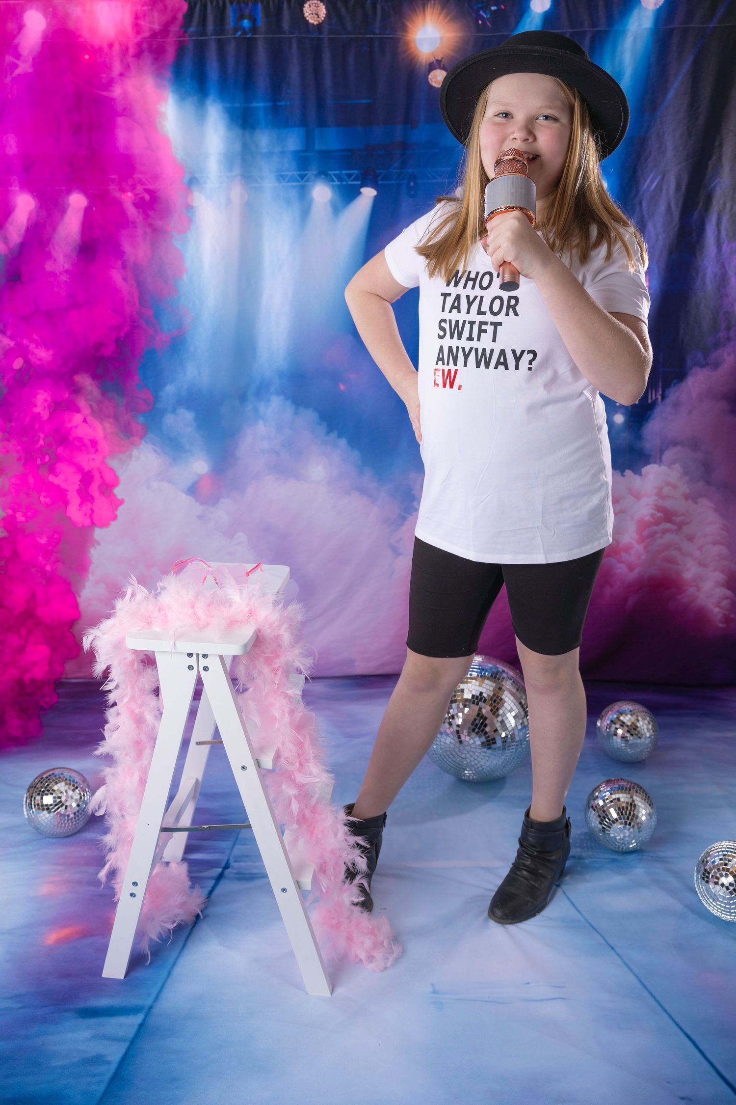 Girl in Who's Taylor Swift Anyway tee and black hat sings into microphone on vibrant pink and blue smoke stage.