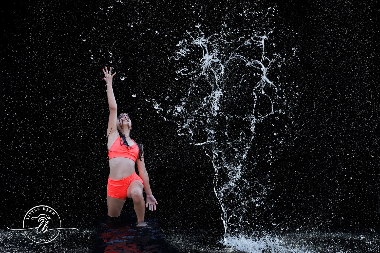 Woman in orange bikini dramatically splashing water against dark background in artistic dance pose.