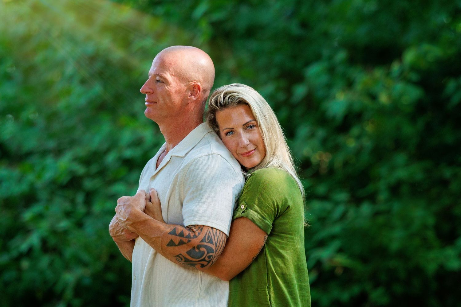 A tattooed bald man and blonde woman pose back-to-back smiling in a bright green outdoor setting.