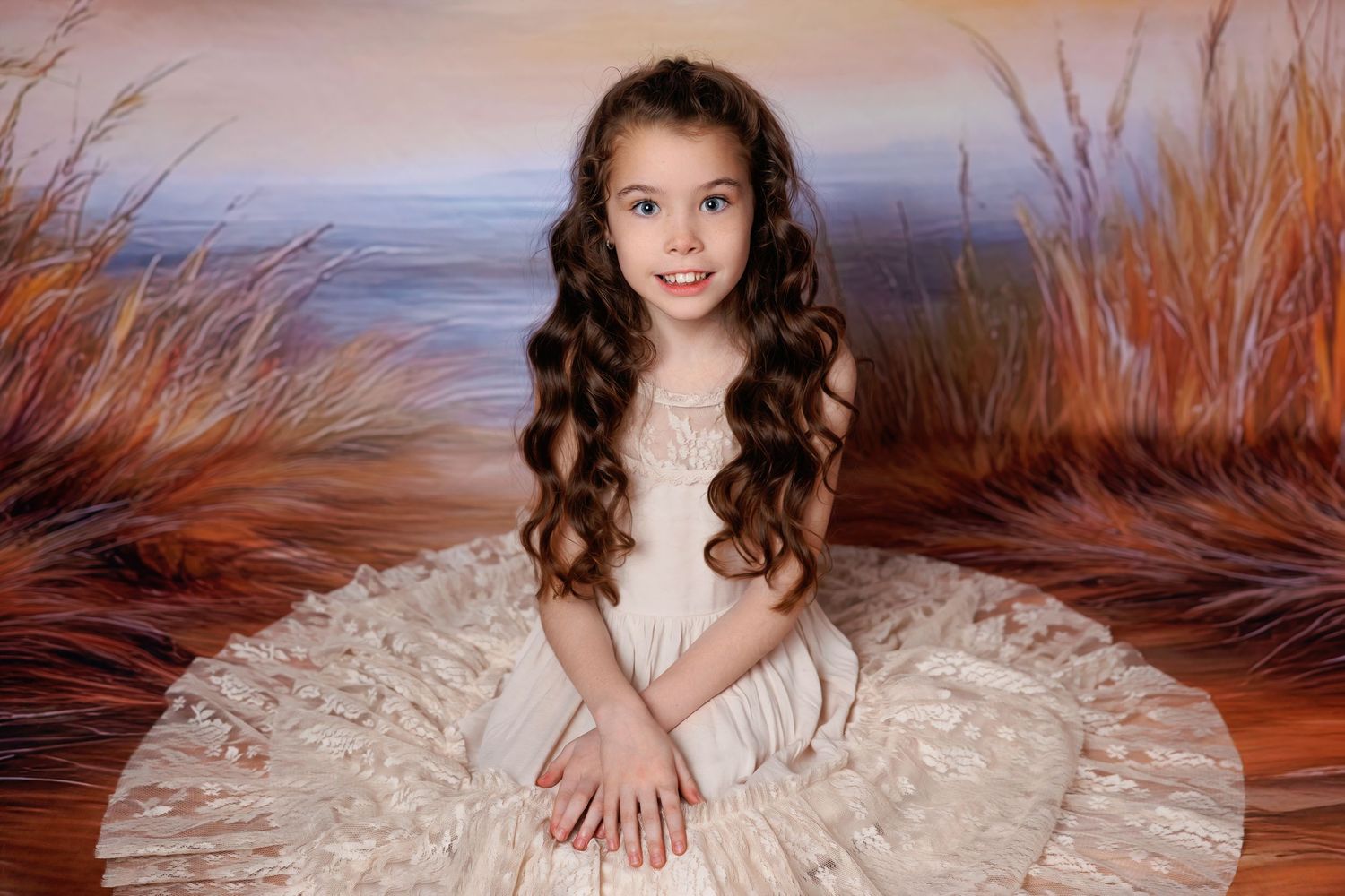 A young girl in a full cream lace gown sits smiling before a painted coastal landscape backdrop.