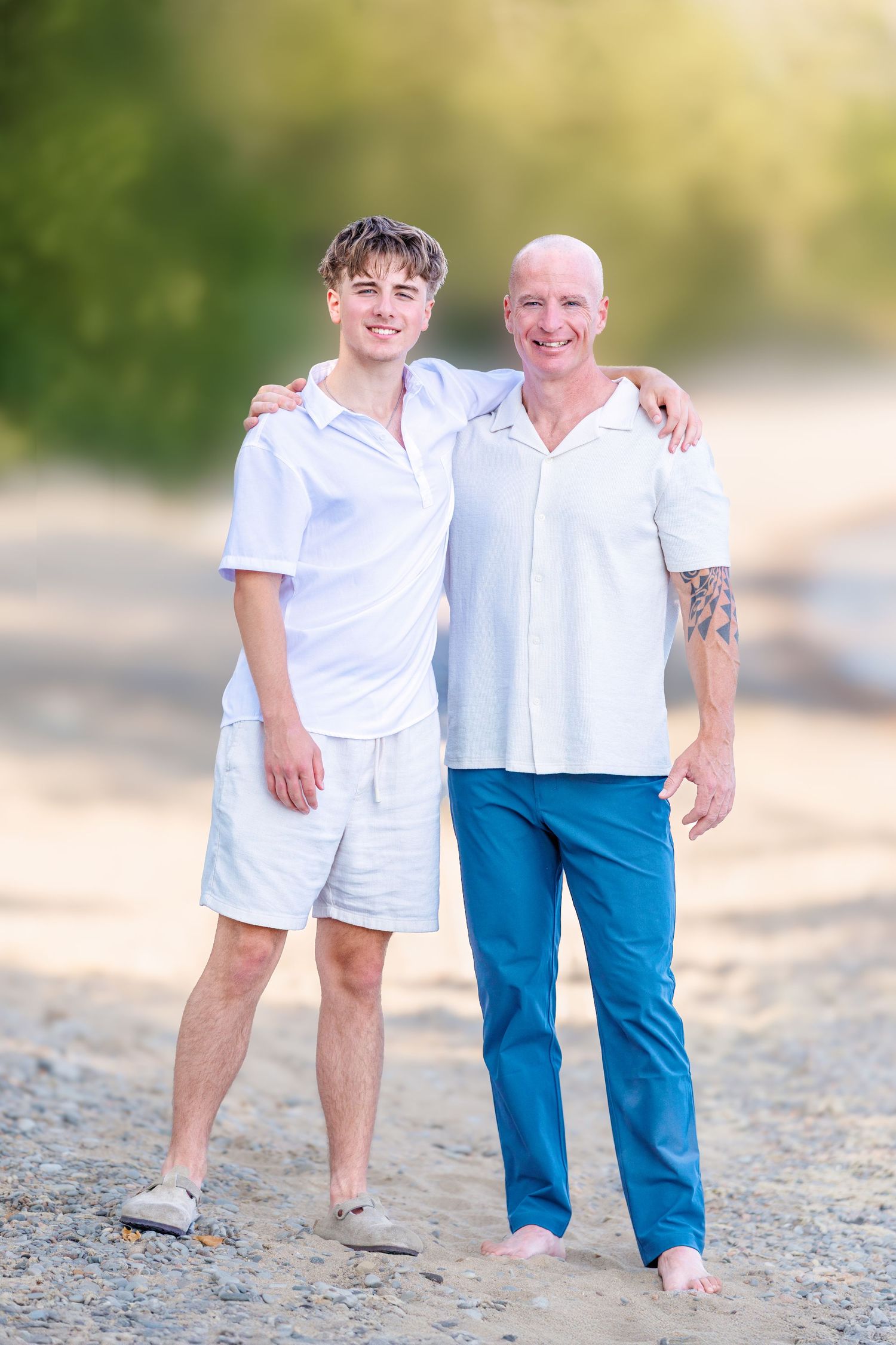 Two males in white shirts embracing on a sandy beach, smiling at the camera in warm natural light.