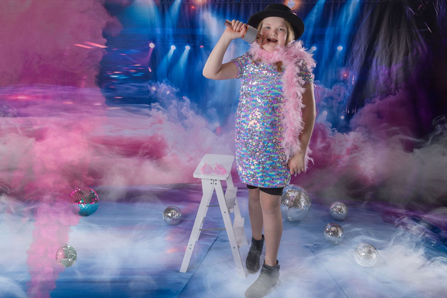 Child performer in sequin dress and feather boa posing on smoky stage with disco balls and spotlights.