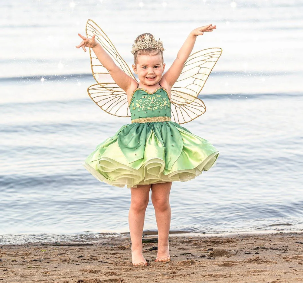 Young girl in green fairy costume with wings and tiara poses joyfully on sandy beach.