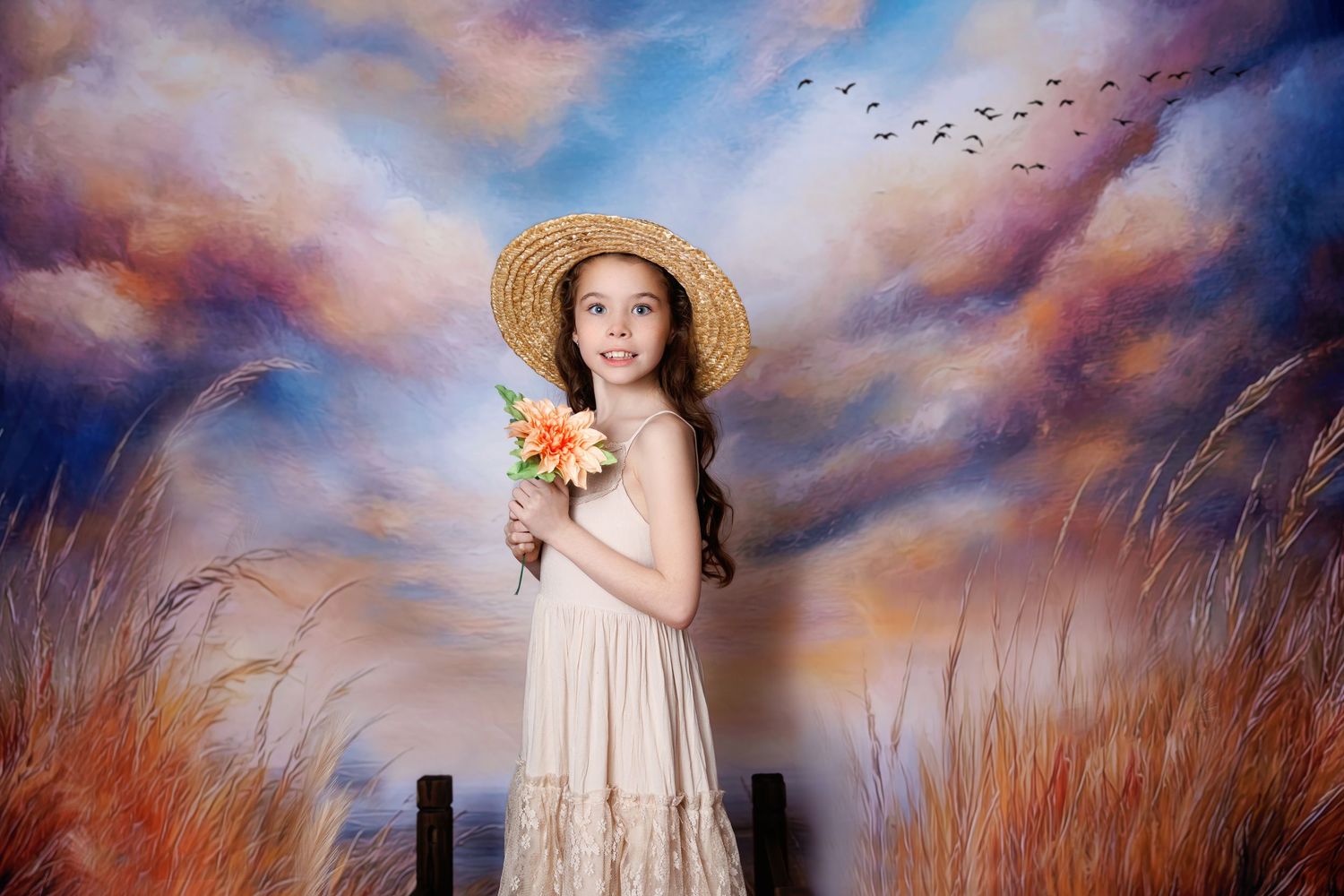A young girl in a straw hat and cream lace dress holds flowers against a painted sunset backdrop.