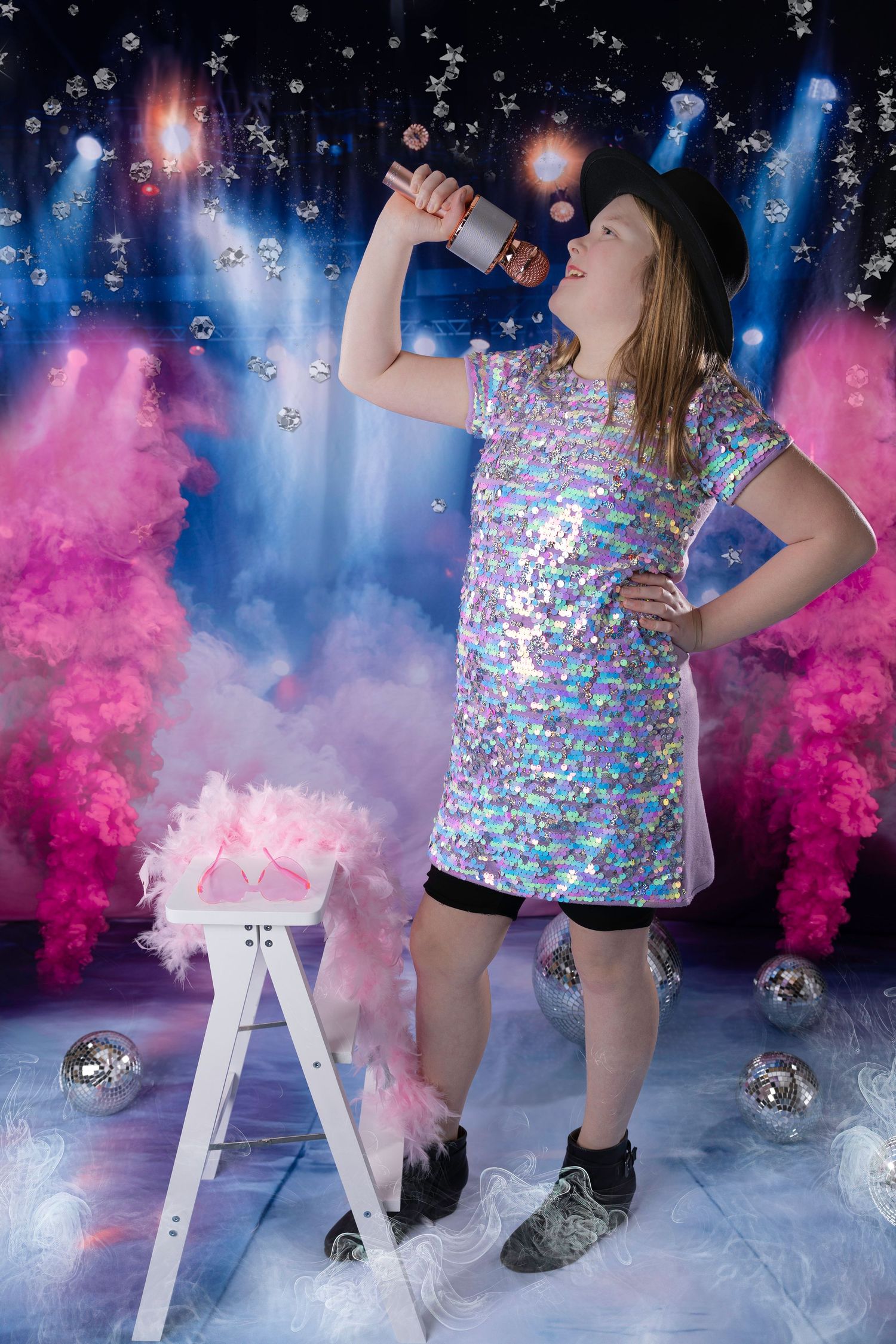 Young girl in iridescent sequin dress and black hat sings into microphone on sparkly pink and blue smoke stage.