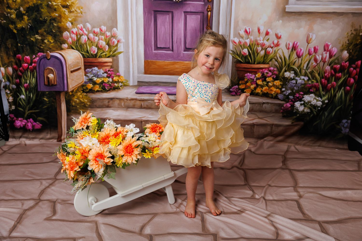 A toddler in a yellow ruffled dress stands beside a floral wheelbarrow in front of a colorful spring backdrop.