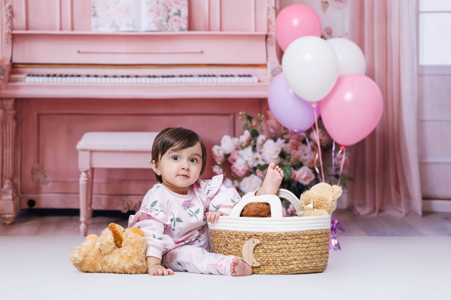A baby girl sits beside a wicker basket with teddy bears and balloons in a pink studio.