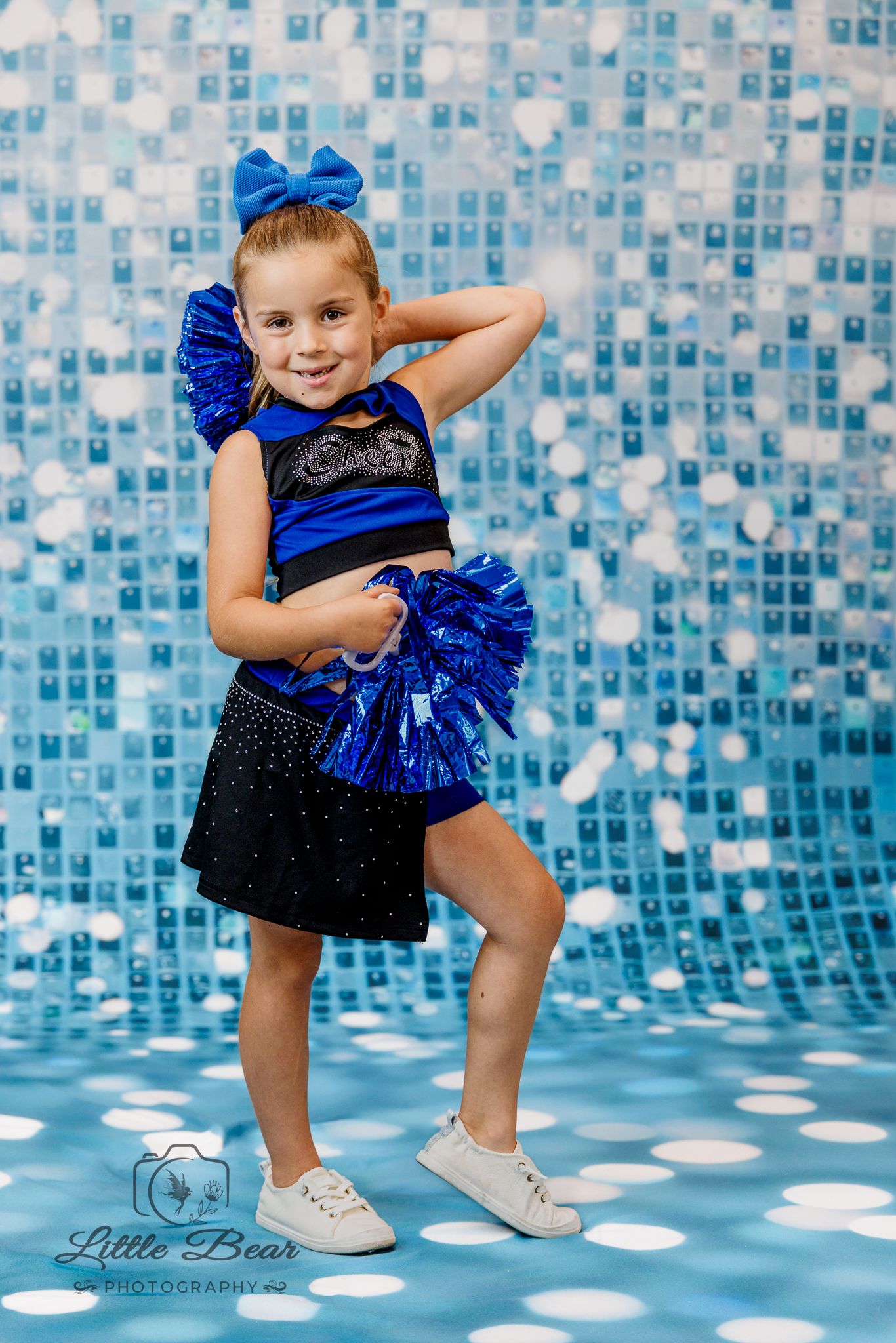 Young girl in blue cheerleader outfit with pom-poms poses confidently against a sparkling blue mosaic backdrop.