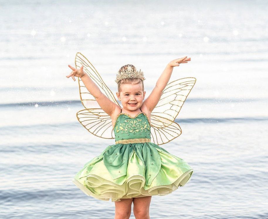 Young girl in green fairy costume with wings and tiara poses joyfully by the water.