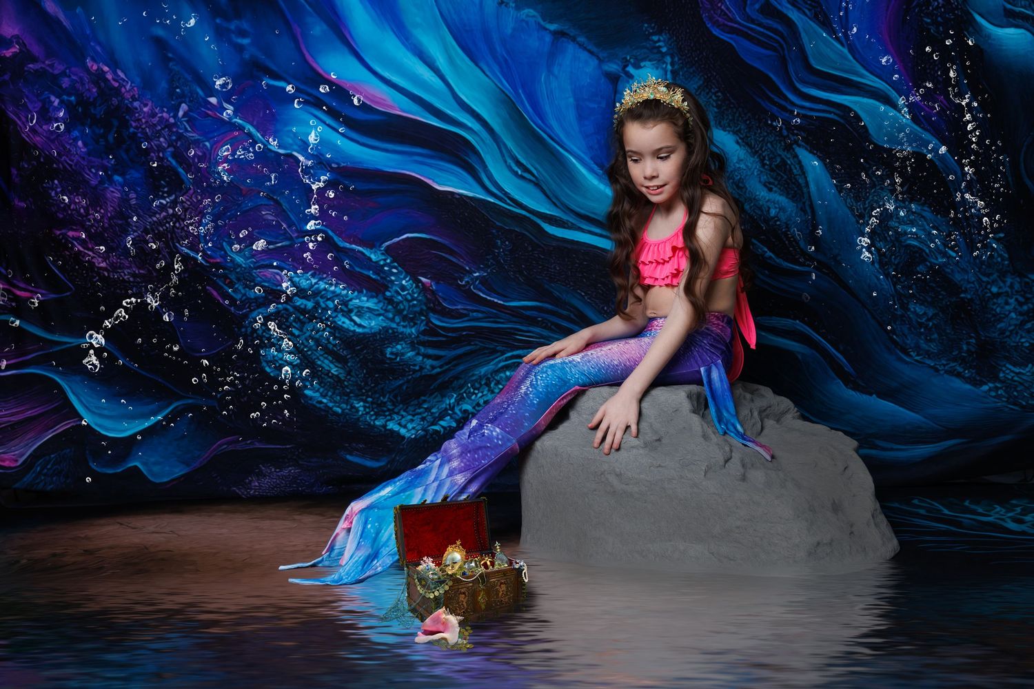A girl dressed as a mermaid with a purple tail and crown sits on a rock near a treasure chest scene.