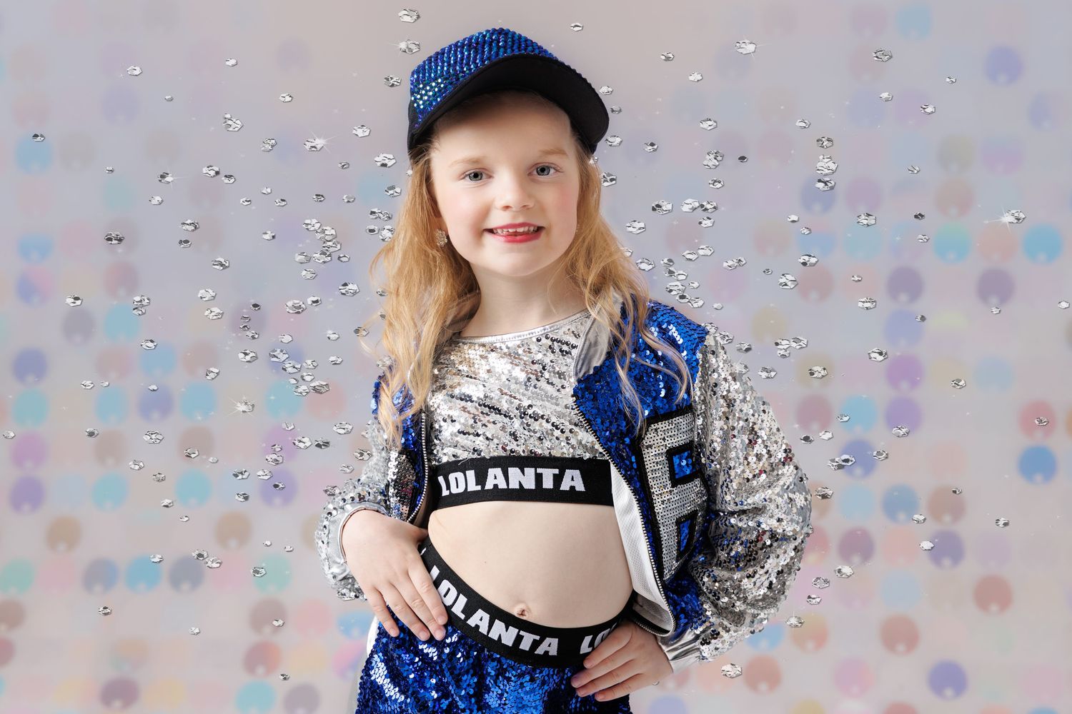 Young girl in sparkly blue sequin dance costume poses against glittery confetti studio backdrop.