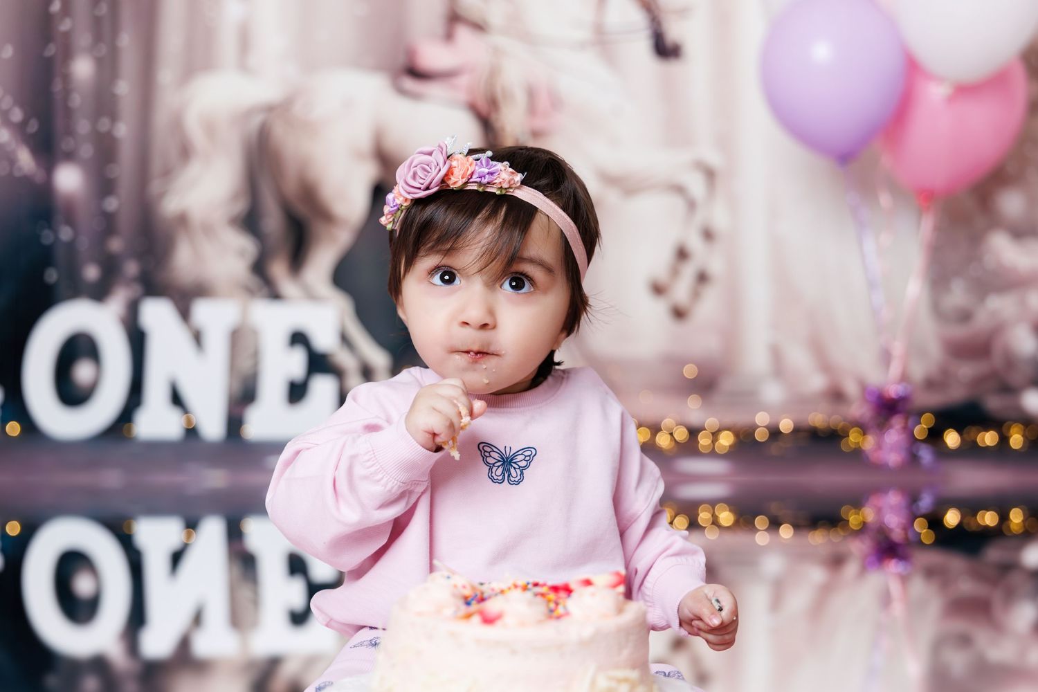 A baby girl in a pink outfit and floral headband reaches toward her first birthday cake.