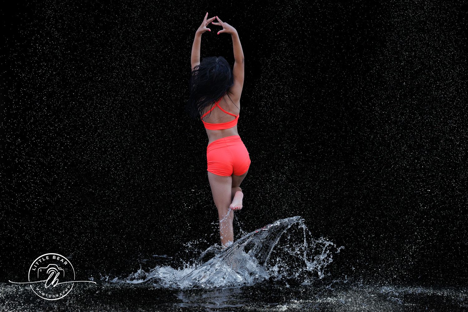 Woman in orange bikini dancing in water splash against dark background.