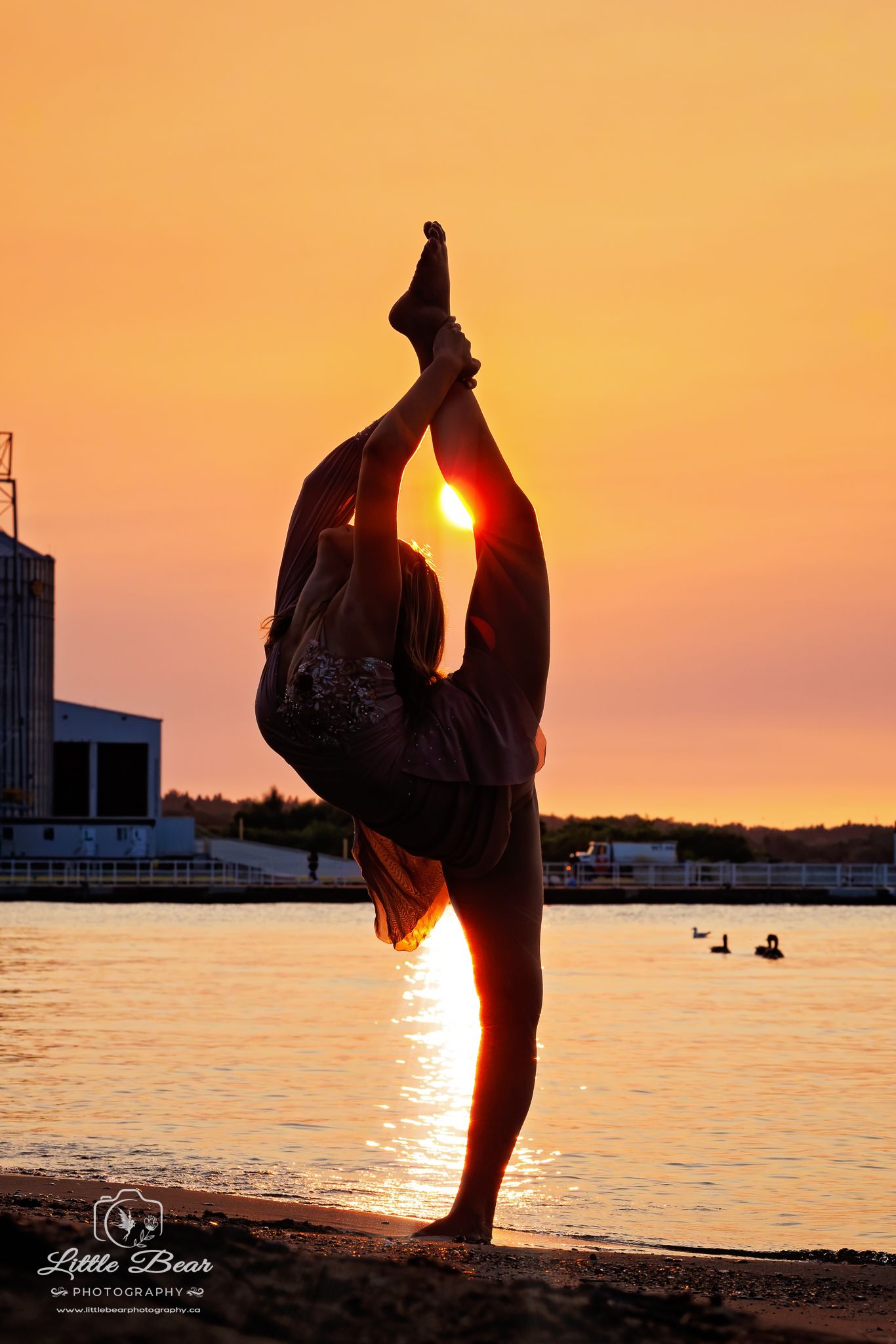 Silhouette of a flexible dancer performing a standing split pose on a beach at golden sunset by the water.
