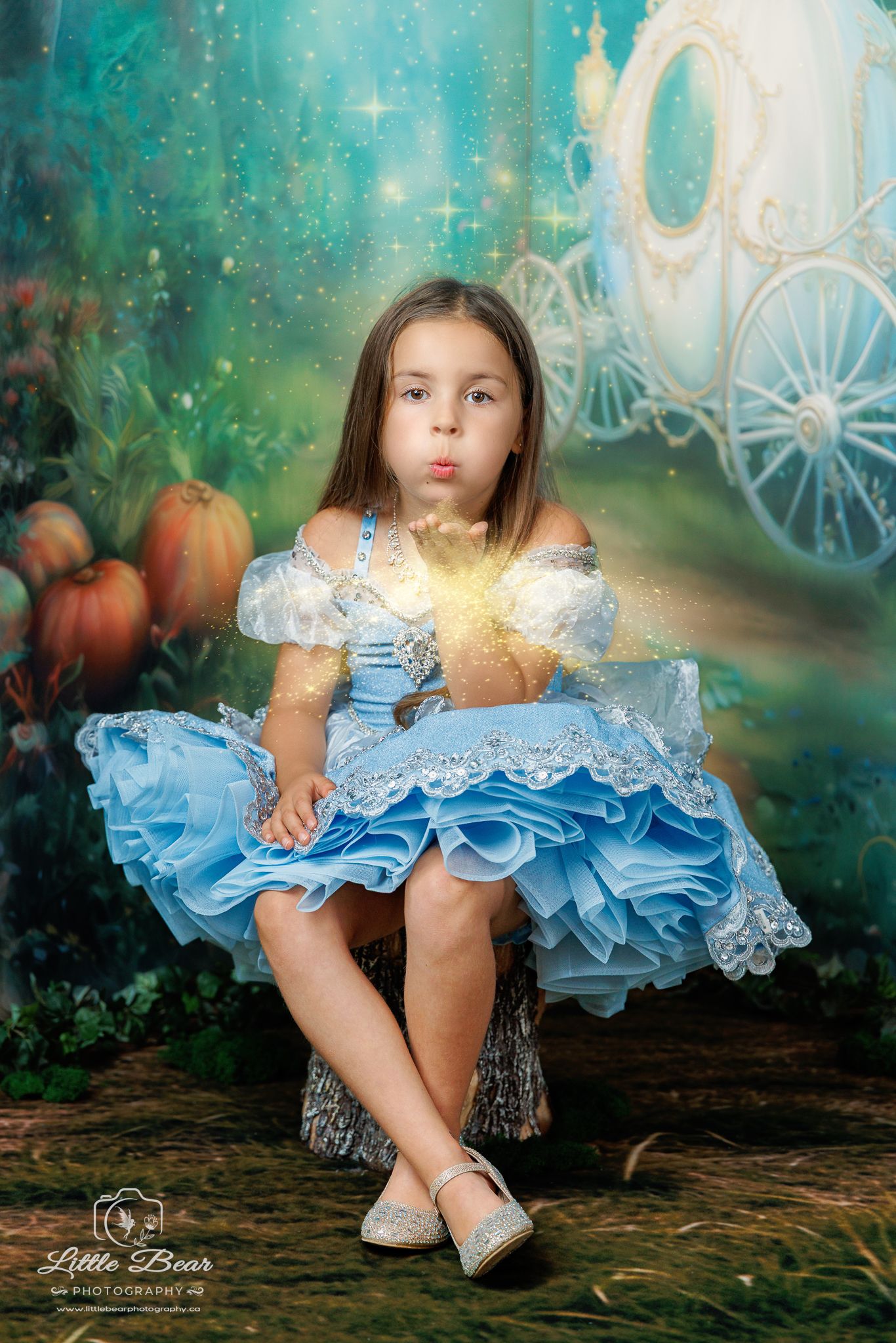A young girl in a blue Cinderella dress blows magical sparkles, posed before an enchanted carriage backdrop.
