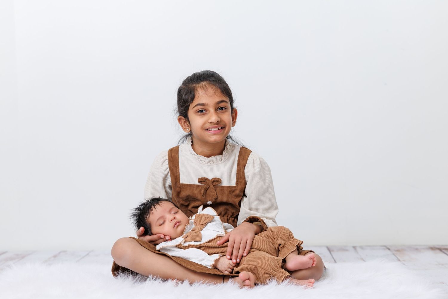 Older child sits cross-legged holding sleeping newborn baby on white fluffy surface.