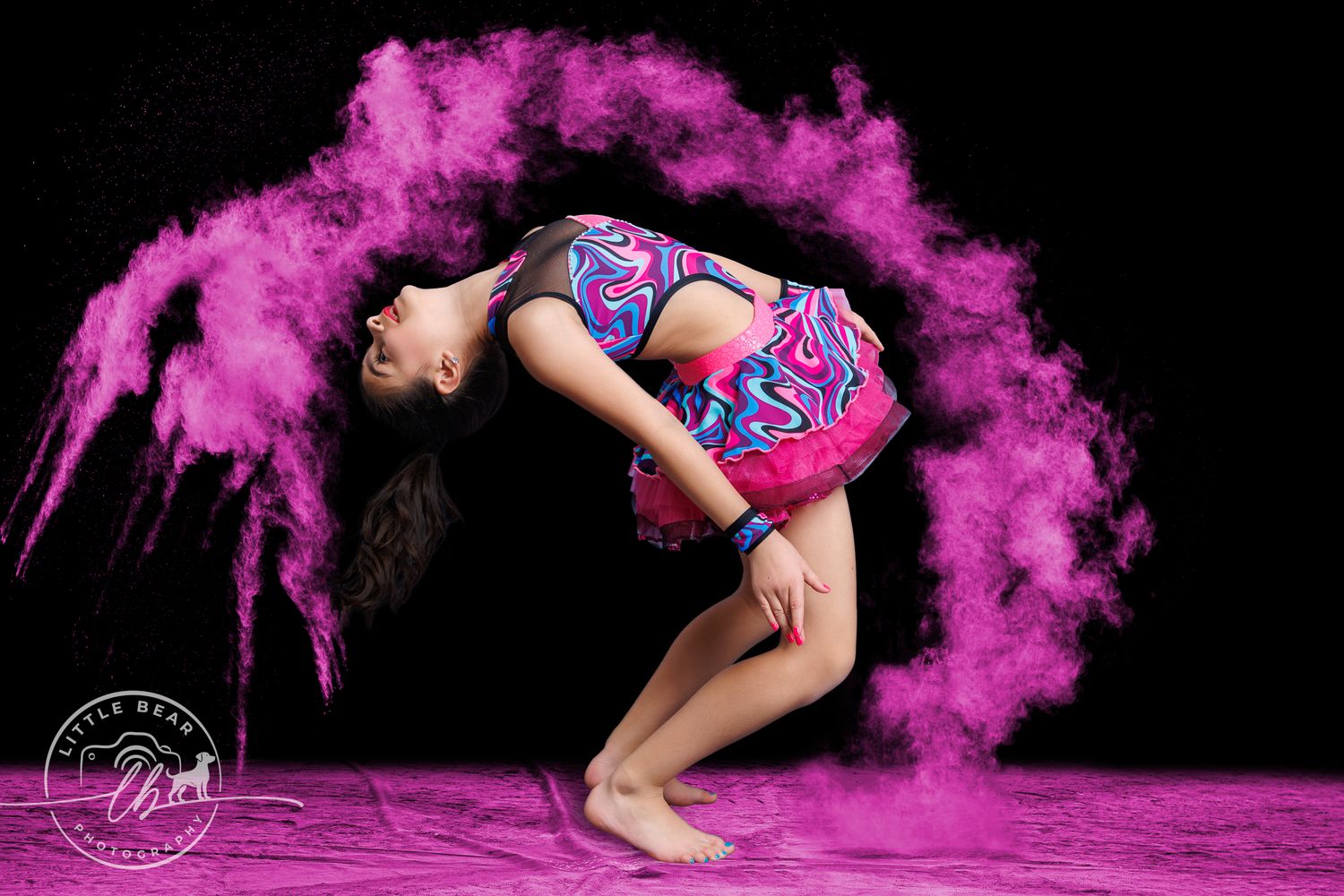 Dancer in vibrant costume poses in backbend surrounded by pink powder explosion.