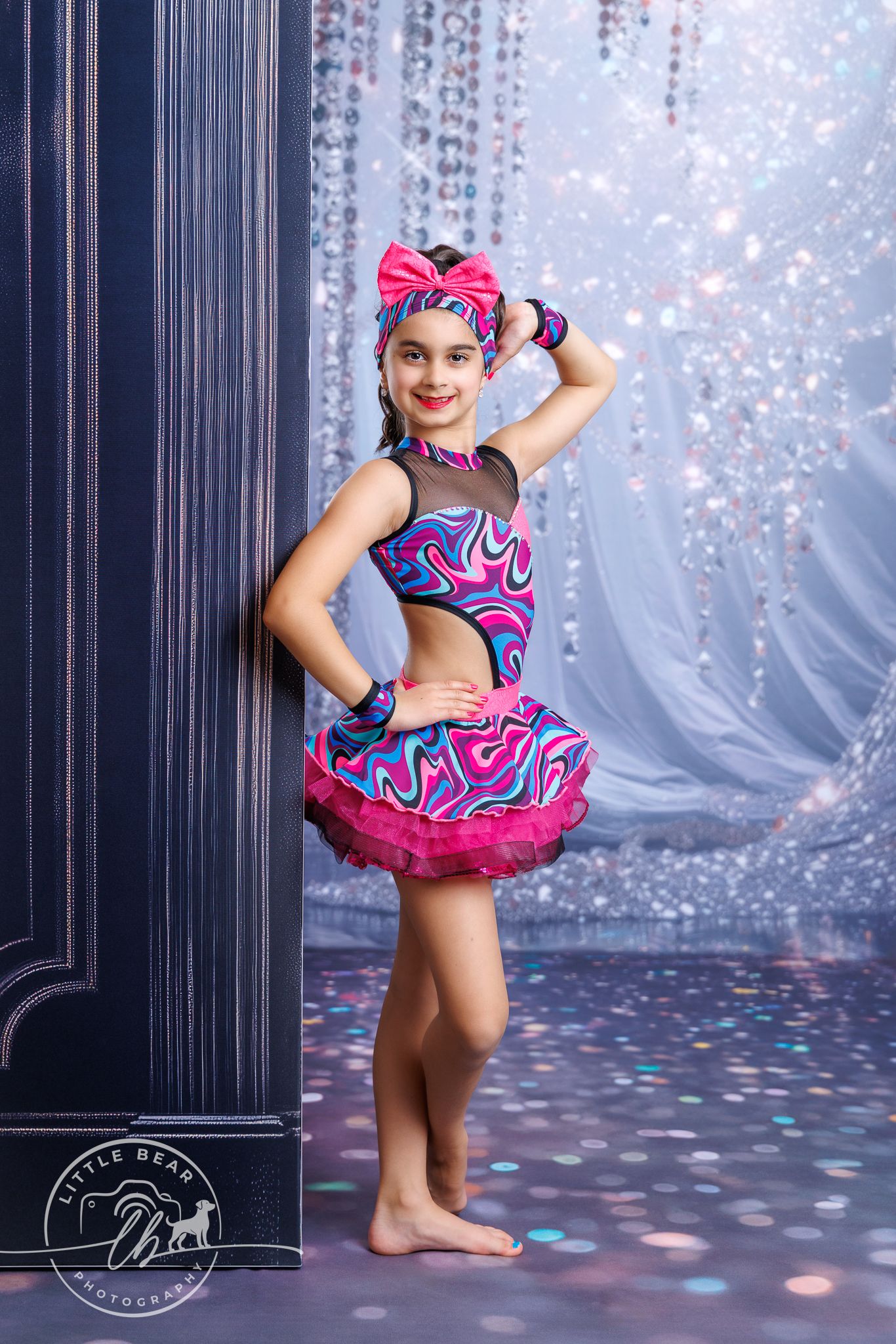 Young dancer in a pink and blue swirl costume poses confidently against a sparkling bokeh backdrop in a professional dance photo.