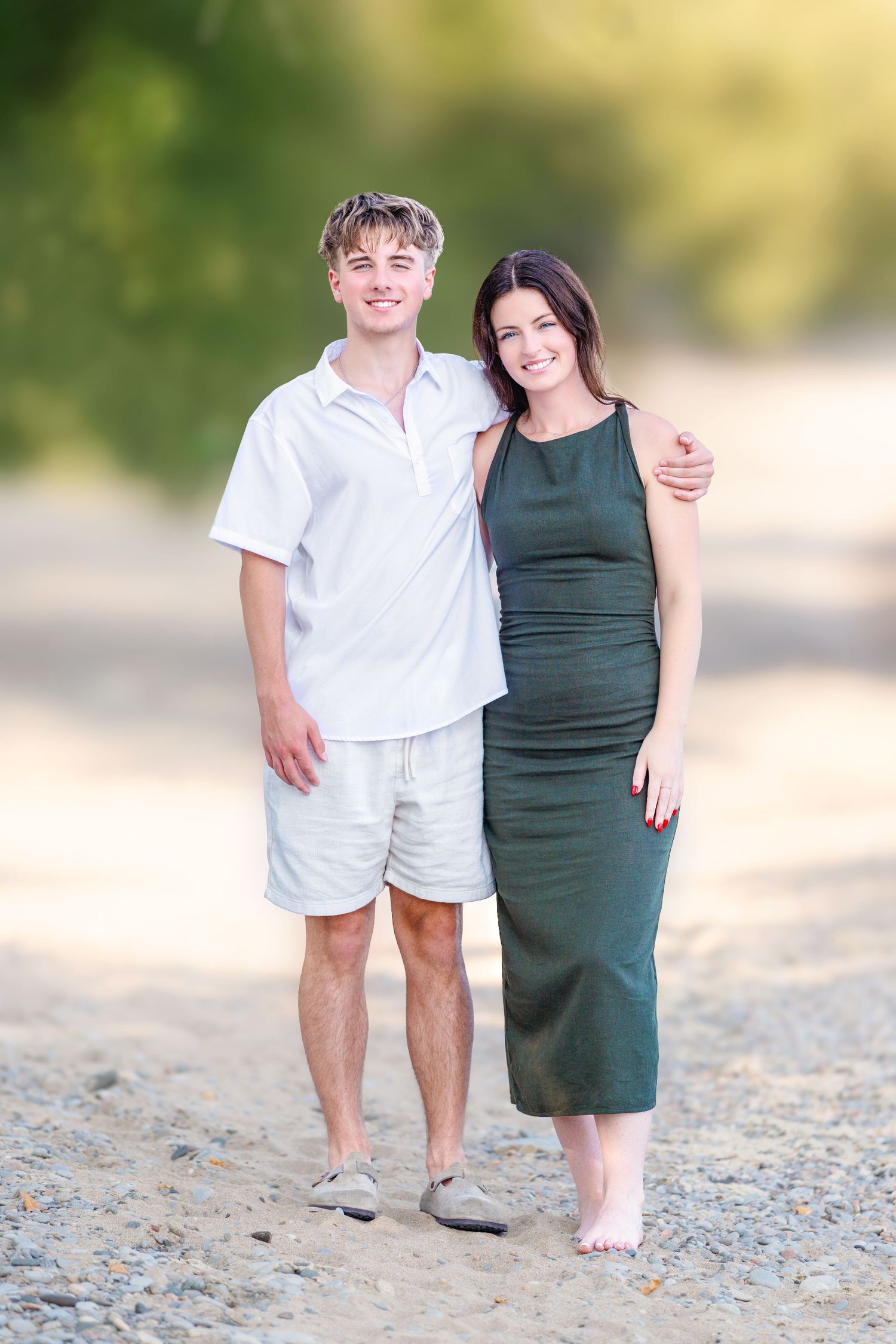 Young man in white shirt and shorts stands with brunette woman in green dress on a sunny beach.