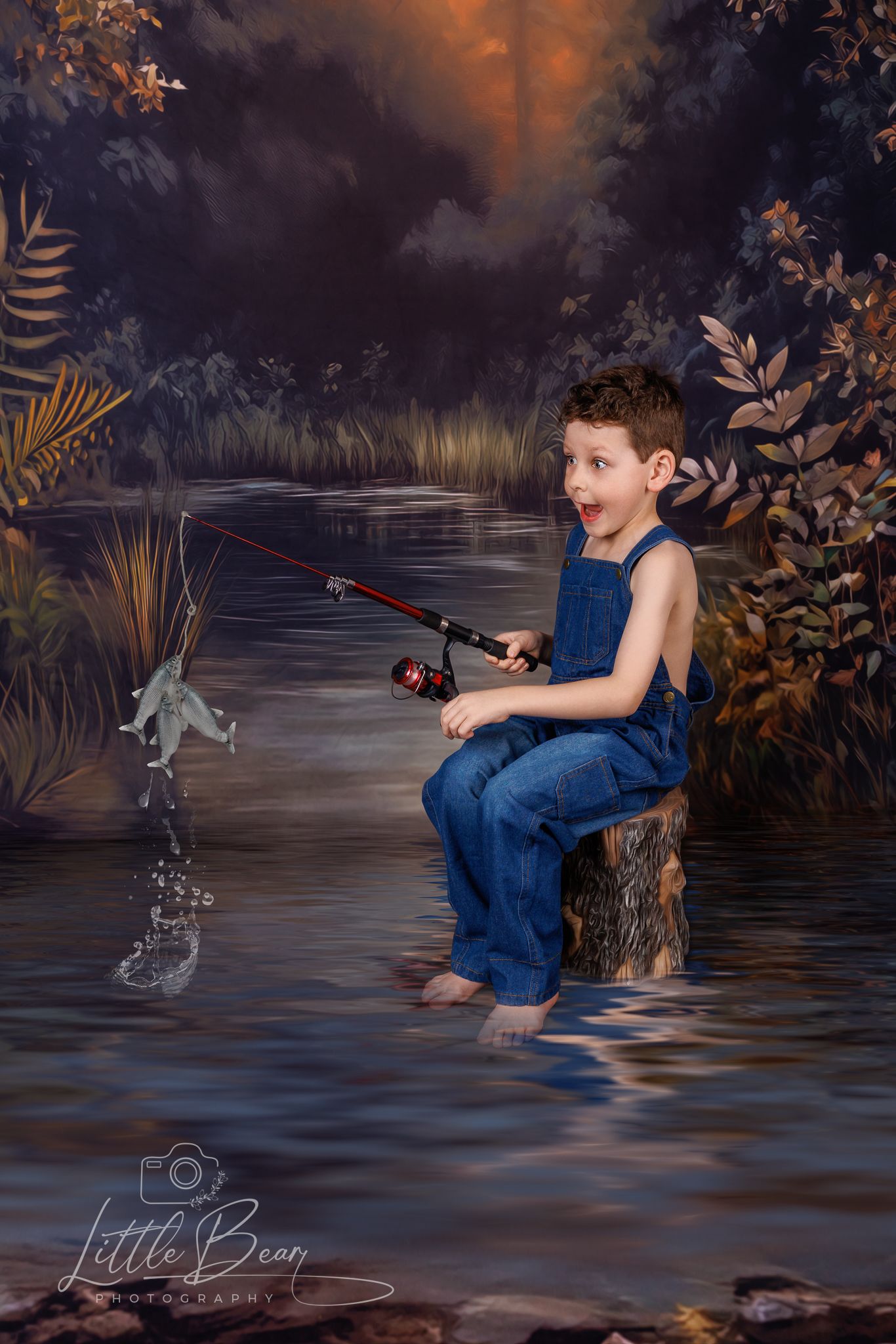 Excited boy in denim overalls catches a fish while sitting on stump in scenic forest water setting.