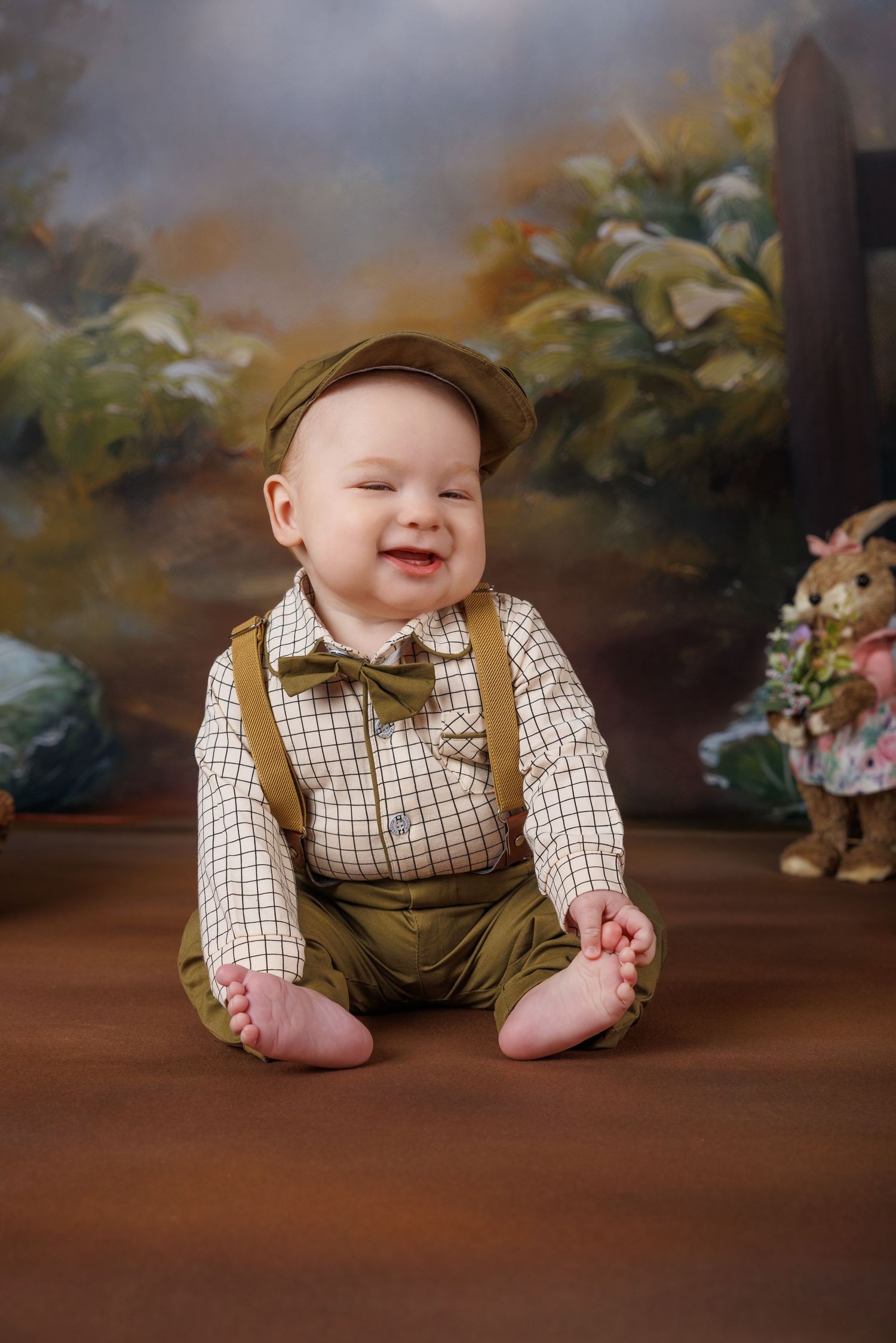 Happy baby boy in vintage green suspenders, bow tie, and cap, smiling against painted backdrop.