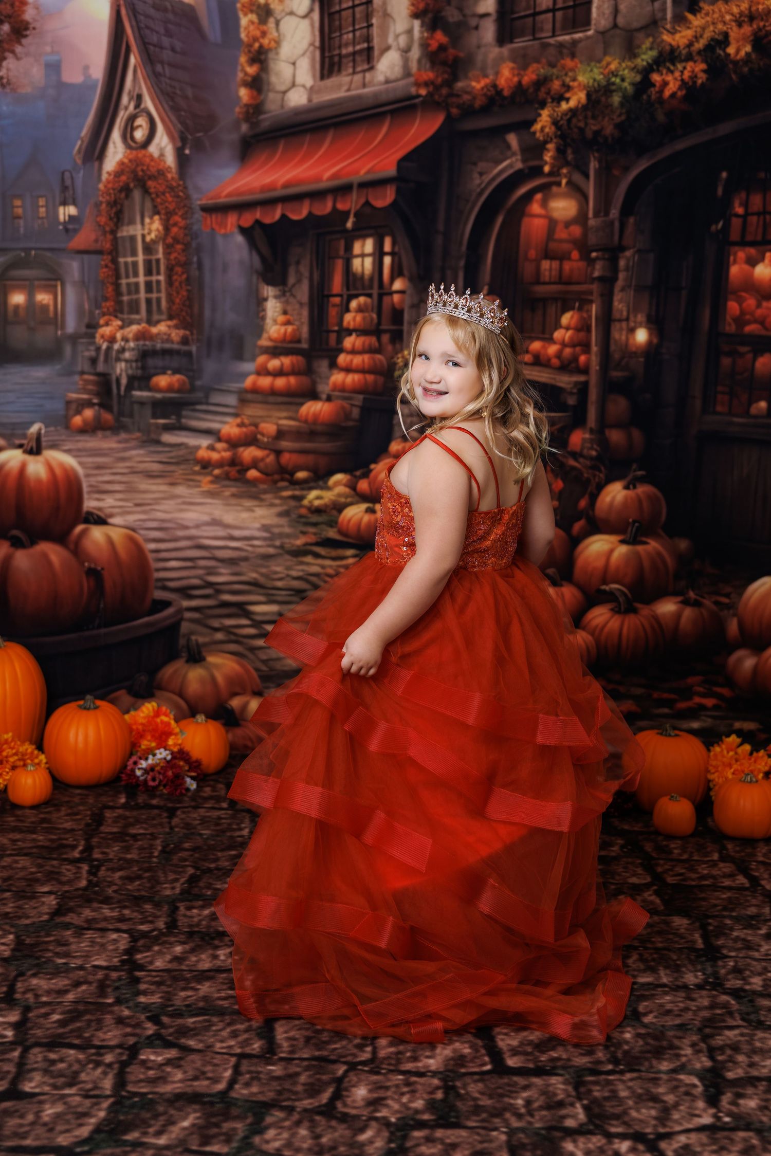 Young girl in red gown and crown poses among pumpkins in a festive autumn village backdrop.