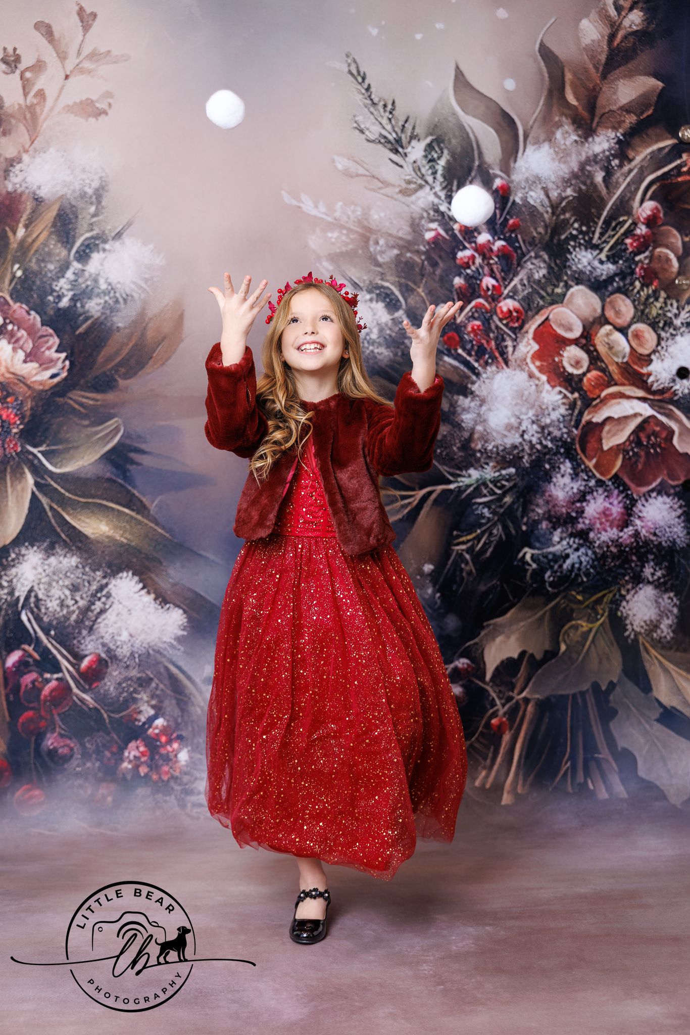 A smiling girl in a red sparkly dress and jacket reaches up joyfully in front of a festive floral winter backdrop.