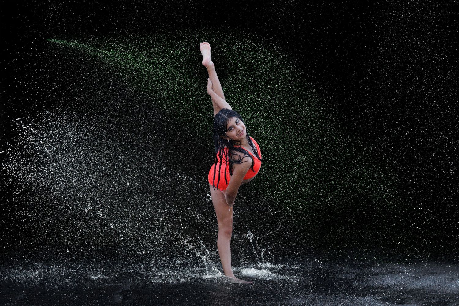 A dancer in a red leotard performs a graceful pose amid dramatic water splashes against a dark background.