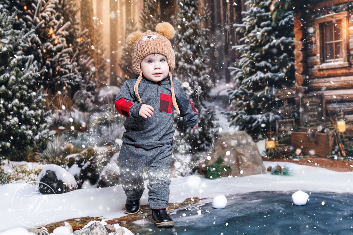 Toddler in winter hat and mittens playing in snow near a cozy log cabin decorated for Christmas.