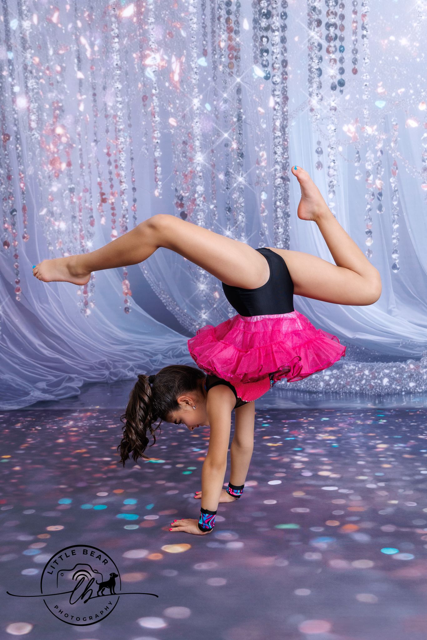 Young dancer performs handstand with legs split in pink tutu on sparkly stage.