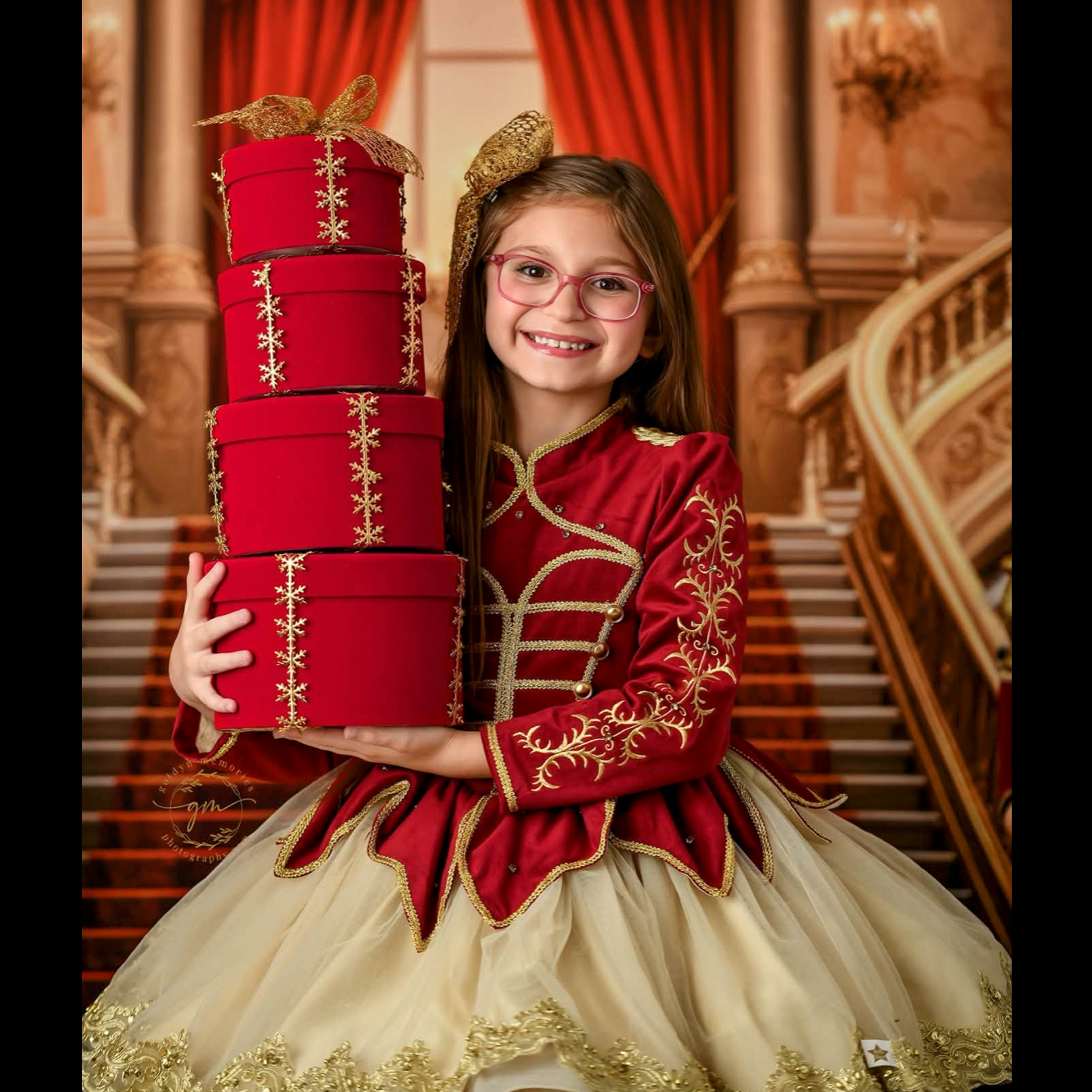 Girl in red and gold nutcracker dress holds stacked holiday gift boxes on grand staircase backdrop.