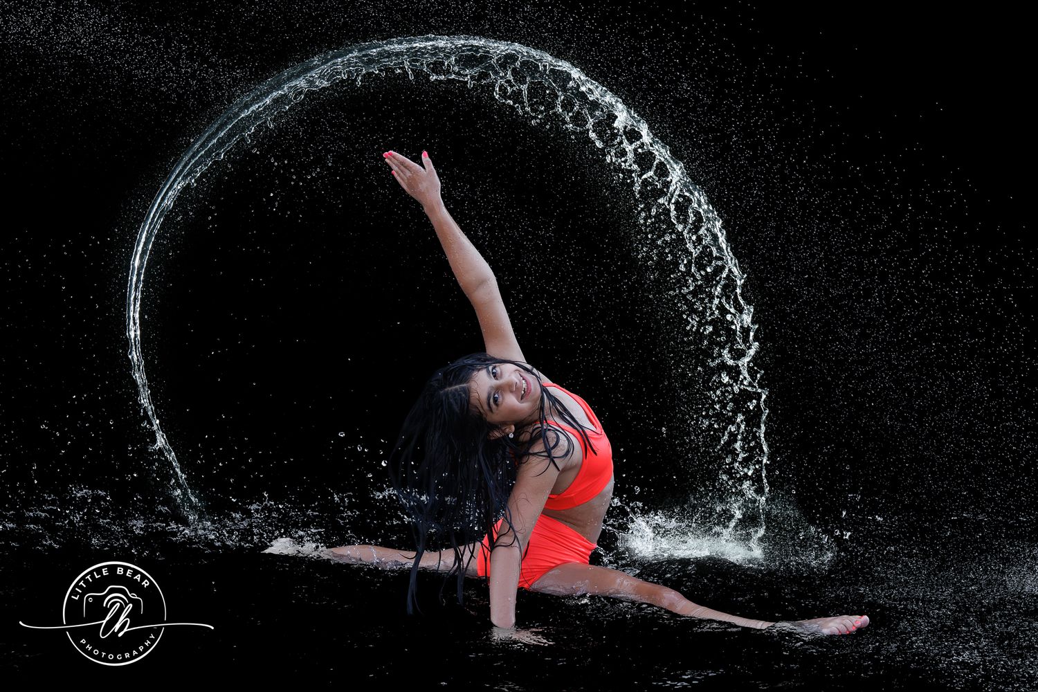 Gymnast in red leotard performs splits while water arcs in a circle behind her against black background.