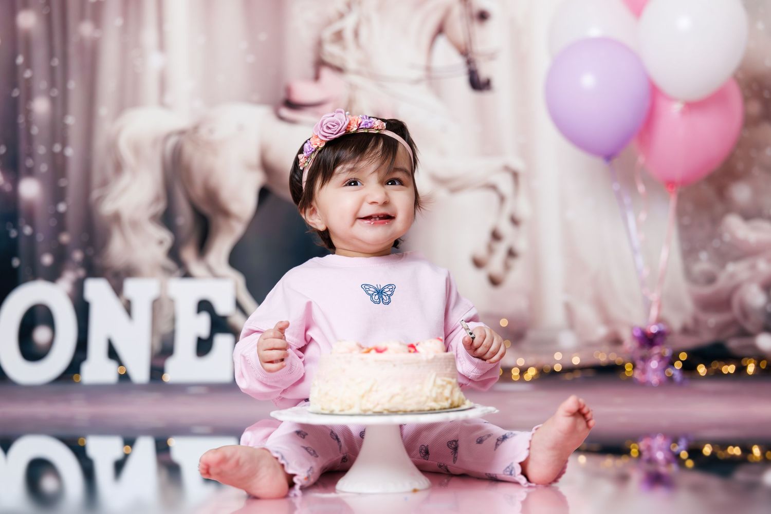 Baby girl in pink outfit smiling at her first birthday cake surrounded by pink balloons and ONE letters.