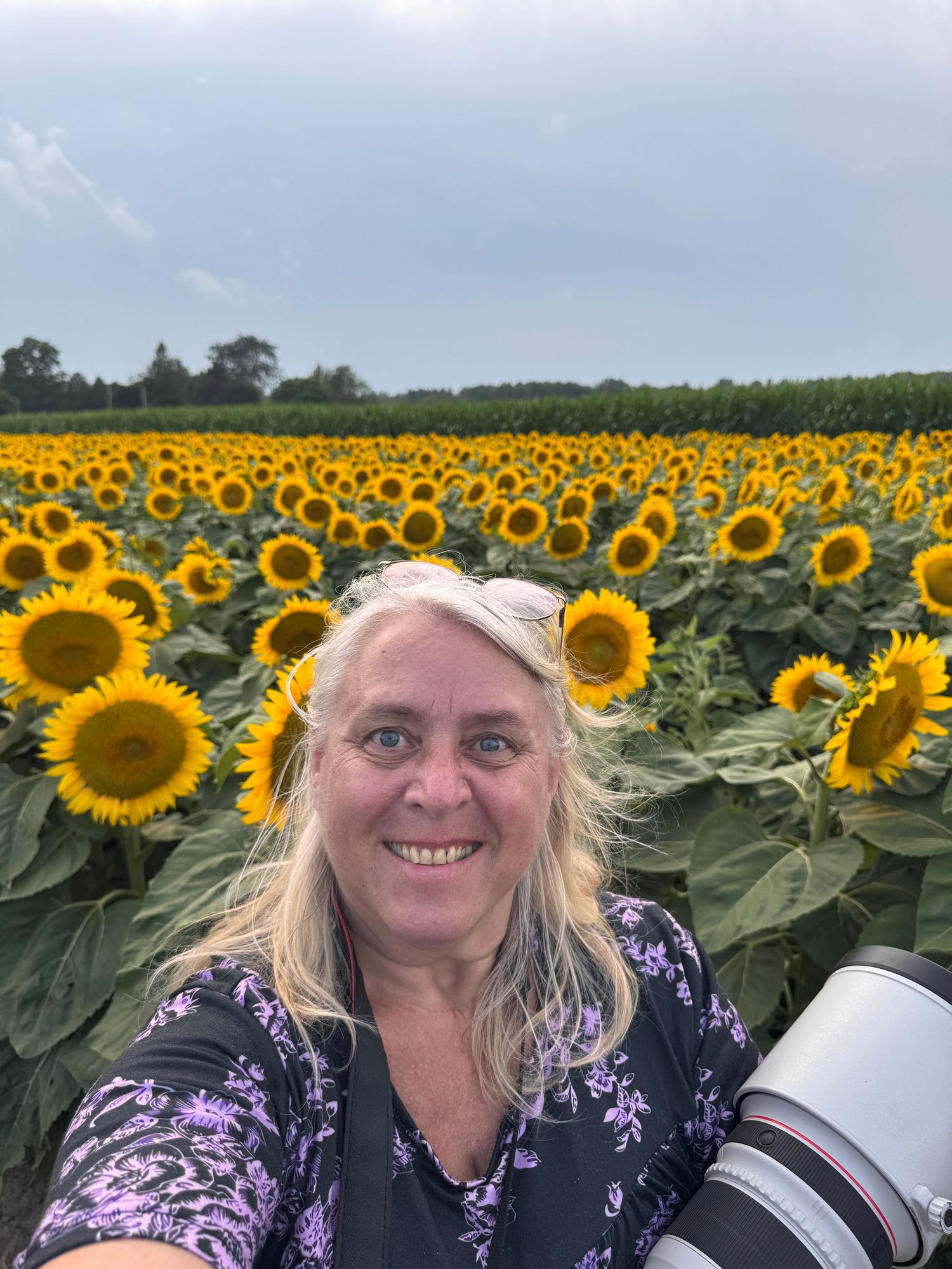 Smiling woman with a camera takes a selfie in front of a vast field of bright yellow sunflowers on a cloudy day.
