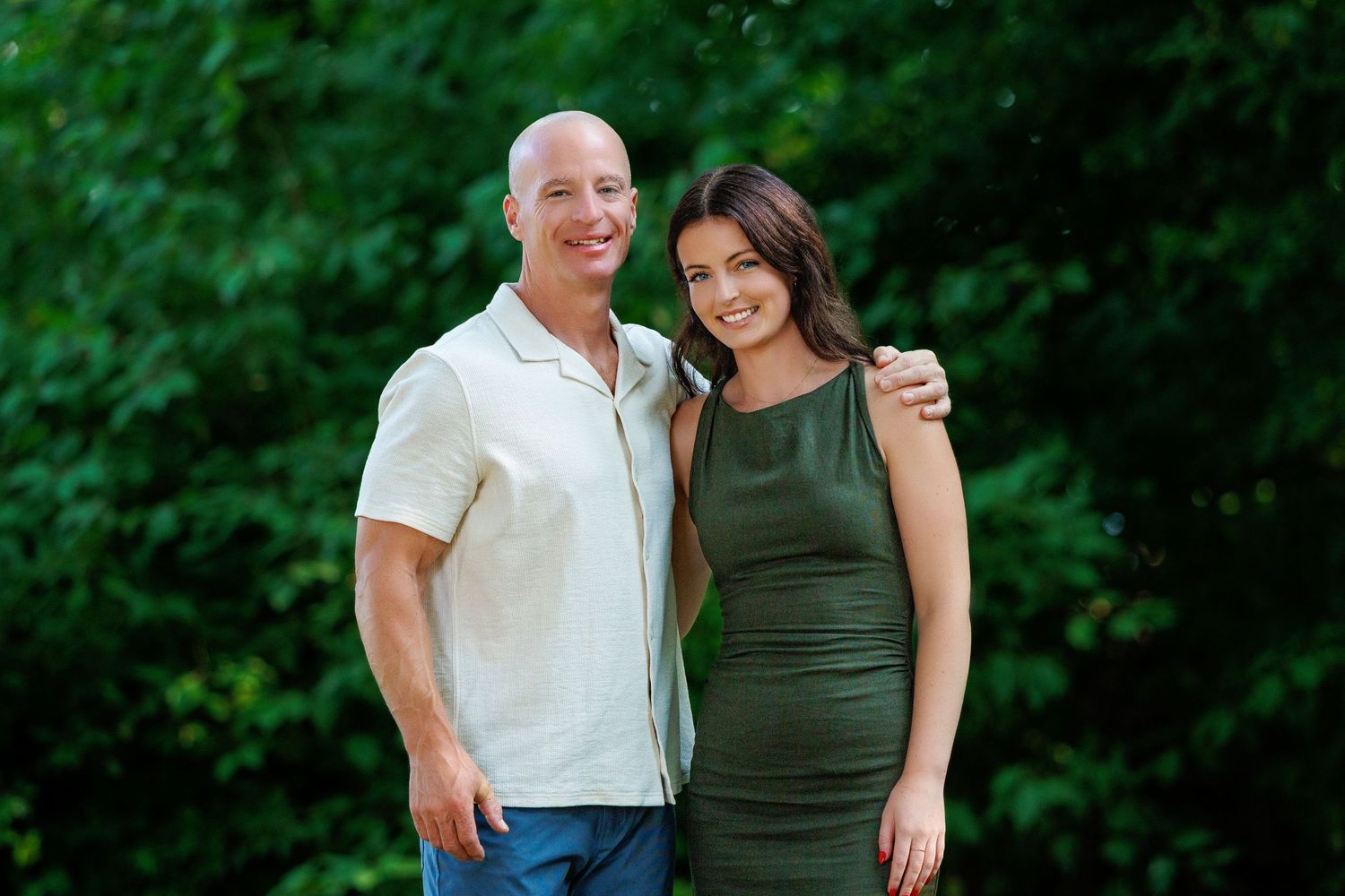 A bald tattooed man poses with a young dark-haired woman in a green dress outdoors.