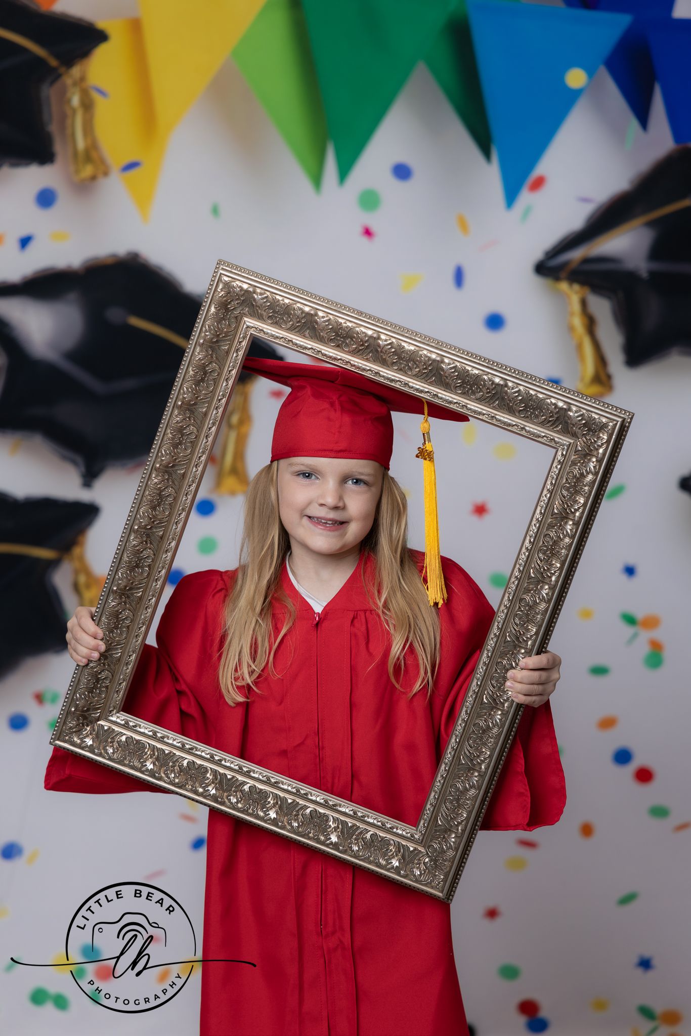 Young girl in red graduation cap and gown holds ornate picture frame, smiling at camera with colorful confetti backdrop.