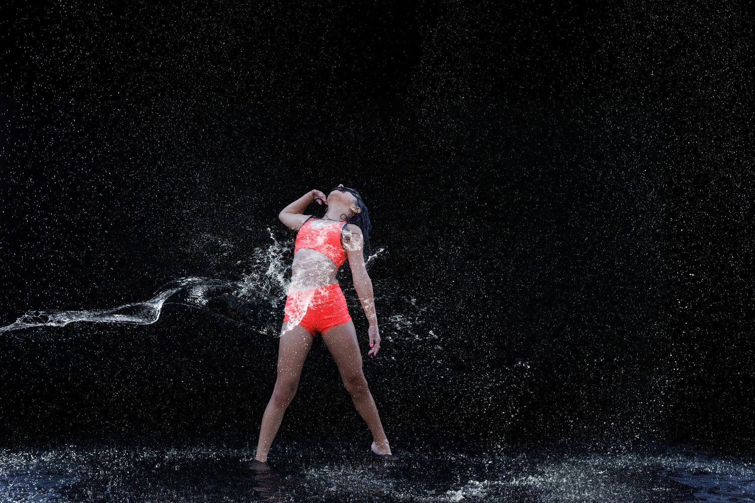 Woman in red bikini dancing in splashing water against dark background.