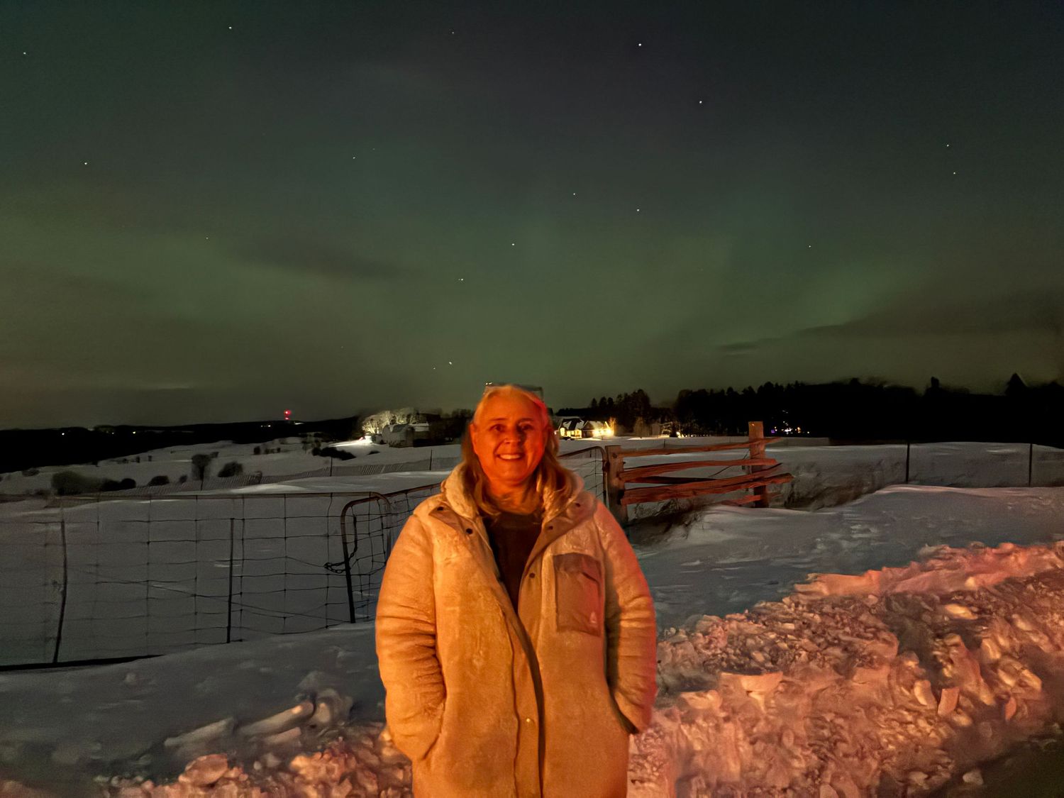 Woman in white winter coat stands outdoors in snowy landscape beneath a vivid green Northern Lights aurora borealis display.