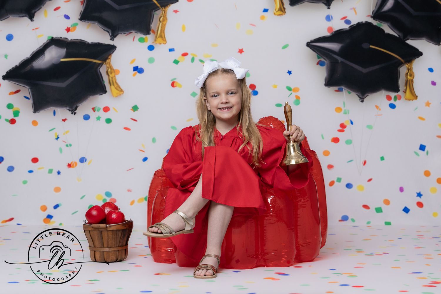 Girl in red graduation gown holds bell, sitting with balloons and confetti in a festive graduation photo.